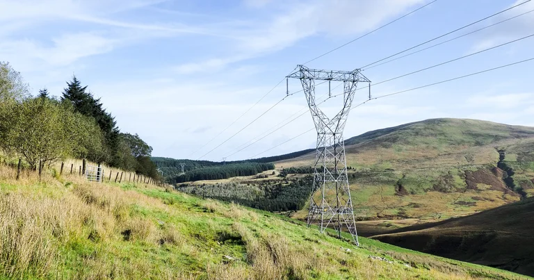 A high-voltage transmission tower carrying power lines across a green hill slope, representative of the Dana-Kushma 220 kV line whose tower in Myagdi was damaged in the September 27 Kaligandaki flood